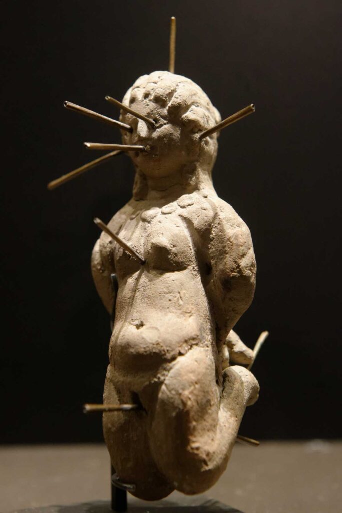 Figurilla de conjuro que representa a una mujer desnuda, arrodillada y atada, atravesada por trece alfileres. Descubierta en un jarrón de terracota que también contenía una tablilla de plomo con una inscripción de defixion (Museo del Louvre).